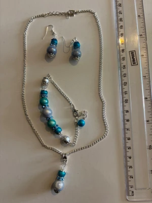 Smyckesset med halsband, örhängen och armband - Snyggt smyckesset med halsband, armband och örhängen i silverfärgad metall med blå, turkosa och silverfärgade pärlor. Kedjorna är tunna och smidiga, och pärlorna har olika nyanser och detaljer. Perfekt för att ge din outfit en färgklick.  Nickelfria 