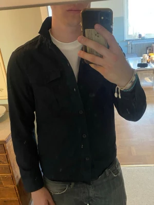 Mörkblå overshirt med bröstficka - Mörkblå overshirt med knäppning framtill och klassisk krage. Modellen har en bröstficka på vänster sida och raka ärmarfrån märket Riley . Perfekt att bära över en t-shirt för en avslappnad look. Materialet ser ut att vara bomull eller en bomullsblandning.