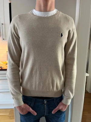 Beige tröja från Ralph Lauren - Stilren beige tröja från Ralph Lauren med rund halsringning och diskret broderad logga i mörkblått på bröstet. Tröjan har långa ärmar och ribbade muddar vid ärmslut och nederkant. Perfekt för en clean och klassisk look.