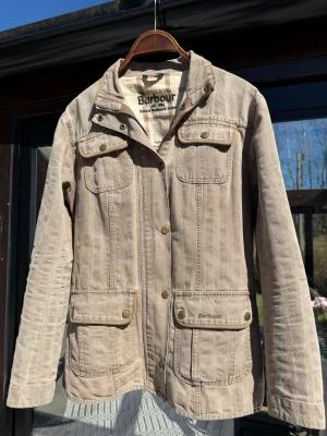 Beige jacka från Barbour - Snygg beige jacka från Barbour med fyra fickor framtill och knappar. Jackan har klassisk krage och lång ärm, perfekt för dig som gillar en stilren och avslappnad look. Tidlös design som funkar till många outfits. Perfekt skick och storlek S. Skriv vid frågor!