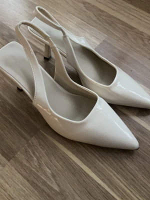 Beiga lackade slingback-pumps - Säljer ett par stilrena pumps i beige lack med spetsig tå och smal klack. Skorna har öppen häl med slingback-rem och en elegant, minimalistisk design. Perfekta för dig som gillar klassiska och trendiga skor med en modern twist.