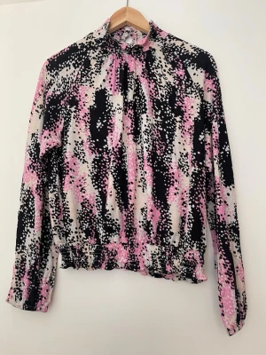 Mönstrad blus med volangkant - Säljer en blus med abstrakt prickigt mönster i svart, rosa och vitt. Blusen har hög krage, långa ärmar och volangdetalj vid midjan. Materialet känns mjukt och stretchigt, perfekt för dig som gillar färg och unika prints.