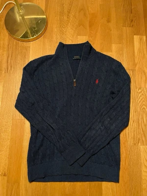 Marinblå stickad tröja Polo Ralph Lauren - Säljer en marinblå stickad tröja från Polo Ralph Lauren med half zip och kabelstickat mönster. Tröjan har långärmad passform, ribbade muddar och röd broderad logga på bröstet. Perfekt för dig som gillar klassisk stil med en modern touch.