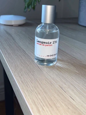 Langmuir 17.0 extrait de parfum - En stilren parfymflaska i genomskinligt glas med silverfärgat lock. Etiketten är vit med svart och röd text där det står Langmuir 17.0 extrait de parfum från one bold chemist. Flaskan har en enkel och modern design.