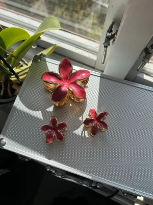 Blommiga hårklämmor i röd och guld - Säljer tre hårklämmor i form av blommor med fem kronblad. Klämmorna är gjorda i metall med röd emalj och guldfärgade kanter. Perfekt för att ge frisyren en färgklick och unik touch.