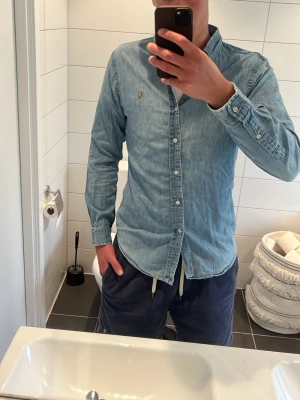 Ljusblå jeansskjorta Polo Ralph Lauren - Snygg ljusblå jeansskjorta från Polo Ralph Lauren i slim fit-modell. Skjortan har klassisk krage, knäppning framtill och lång ärm. Ikonisk broderad logga på bröstet. Perfekt för en avslappnad men stilren look. Priset är inte hugget i sten så kom gärna med prisförslag. Som ni ser på sista bilden så har den ett litet vilket inte syns så tydligt när man har på den eftersom det är i armhålan. Modellen är 187cm och väger 80 kg