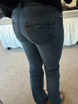 Mörkblå bootcut jeans med fickor - Säljer ett par mörkblå bootcut vintage jeans med klassiska fem fickor och diskreta slitningar. Jeansen har låg midja och raka ben med lätt utsvängning nertill. Perfekta för en avslappnad och trendig look.