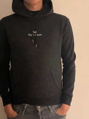 Hoodie från Polo Ralph Lauren - Snygg mörkgrå/svart hoodie från Polo Ralph Lauren med broderad logga på bröstet och ärmen. Klassisk huva med dragsko och känguruficka framtill. Mjuk bomullsblandning som är skön att ha på sig. Perfekt för en avslappnad och stilren look.