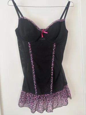 Svart babydoll med leoparddetaljer - Svart babydoll med rosa leopardmönster och matchande trosa. Babydollen har justerbara axelband och en liten rosett framtill. Trosan har också en liten rosett framtill. 