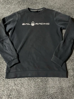 Svart Sail Racing sweatshirt - Svart sweatshirt från Sail Racing med vit logotyp och text på bröstet. Tröjan har rund hals, långa ärmar och ribbade muddar. Materialet är mjukt och ger en sportig vibe. Perfekt för dig som gillar stilrena och bekväma plagg.