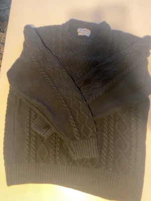 Svart kabelstickad tröja New Garments - Säljer en svart kabelstickad tröja från New Garments i storlek L. Tröjan har ett klassiskt flätmönster, rund halsringning och ribbade muddar vid ärmslut och nederkant. Perfekt för dig som gillar tidlös och enkel stil.