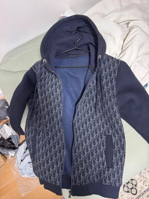 Mörkblå hoodie med mönster - Snygg mörkblå hoodie med dragkedja och huva. Framsidan har ett stilrent mönster i grått och ärmarna är enfärgade. Hoodien har fickor på sidorna och ribbade muddar. Perfekt för en chill och trendig look.