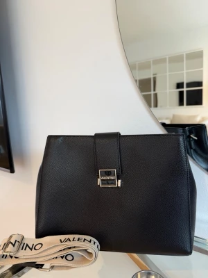 Svart väska från Valentino - Stilren svart axelväska från Valentino i slätt läder med silverfärgat spänne framtill. Väskan har en avtagbar och justerbar axelrem i beige med svart logotyp. Perfekt för dig som gillar klassisk och modern design.
