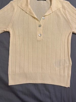 Beige ribbad topp från Gina Tricot - Snygg beige ribbad topp från Gina Tricot med korta ärmar och krage. Toppen har tre knappar framtill och är tillverkad i ett mjukt, stickat material. Perfekt för en trendig och avslappnad look.