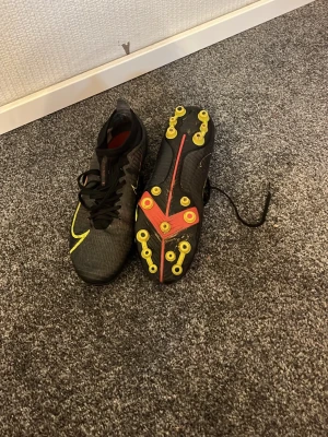 Nike svarta fotbollsskor - Säljer ett par Nike fotbollsskor i svart med gula och röda detaljer. Skorna har snörning, låg profil och mönstrad ovandel för bra bollkontroll. Sulan har fasta dobbar i gult för optimalt grepp på gräsplan. Perfekta för dig som vill ha stil och prestanda på planen.