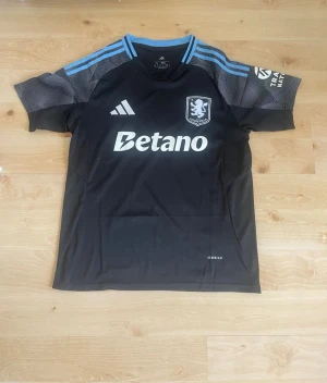 Aston Villa svart matchtröja Adidas 25/26 - Snygg svart Aston Villa matchtröja från Adidas med ljusblå detaljer på axlarna och klubbmärke på bröstet. Tröjan har Betano-tryck framtill och meshpaneler på ärmarna för extra ventilation. Perfekt för fotbollsträning eller att bära som supporter.