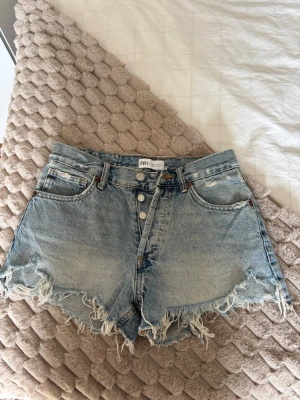 Blåa denimshorts från Zara - Säljer ett par blåa denimshorts från Zara med hög midja och råa, fransiga kanter. Shortsen har knappar framtill, fem fickor och bälteshällor. Perfekta för sommaren och ger en avslappnad vibe.