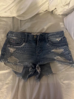 Hollister shorts  - Säljer ett par blå jeansshorts från Hollister med slitna detaljer, råa kanter och små silverfärgade nitar framtill. Shortsen har låg midja och klassisk femficksdesign. Perfekta för sommaren och ger en cool, avslappnad vibe.