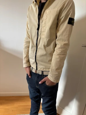 Beige overshirt från Stone Island - Snygg beige overshirt från Stone Island med klassisk krage, svart dragkedja och bröstficka. Ikonisk logotyp-patch på vänster ärm. Långärmad och tillverkad i bomull, perfekt för lager-på-lager-stil.