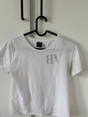 Vit t-shirt med strasstext - Vit t-shirt från Bik Bok med korta ärmar och rund hals. På bröstet står det 'VIP' i strass och på ryggen 'NEED MONEY FOR PORSCHE' i samma stil. T-shirten har en croppad passform och är gjord i mjuk bomull.
