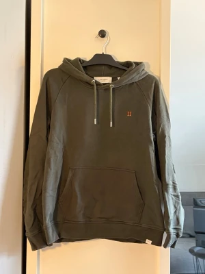 Olivgrön hoodie Les Deux - Snygg olivgrön hoodie från Les Deux med huva. Klassisk känguruficka framtill och diskret broderad detalj med romerska siffran II på bröstet. Tillverkad i mjuk bomull, perfekt för en avslappnad stil.