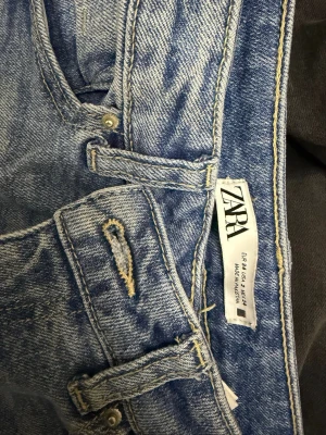 Blå raka jeans från ZARA - Ett par klassiska blå jeans från ZARA med rak passform och hög midja. Jeansen har fem fickor, bälteshällor och stängs med knapp och dragkedja. Perfekta för en avslappnad och trendig look.