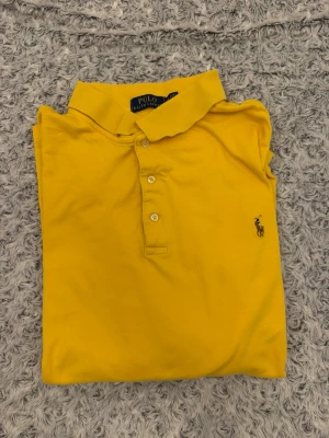 Gul pikétröja från Ralph Lauren - Snygg gul pikétröja från Ralph Lauren med klassisk krage och tre knappar framtill. Tröjan har det ikoniska broderade logotypen på bröstet och är tillverkad i mjuk bomull. Perfekt för en fräsch och sportig look.