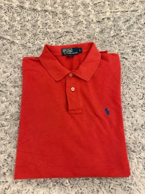 Röd pikétröja från Ralph Lauren i strl L - Klassisk röd pikétröja från Polo Ralph Lauren med blå broderad logga på bröstet. Tröjan har krage, knappar framtill och är tillverkad i bomull. Perfekt för en stilren och avslappnad look.