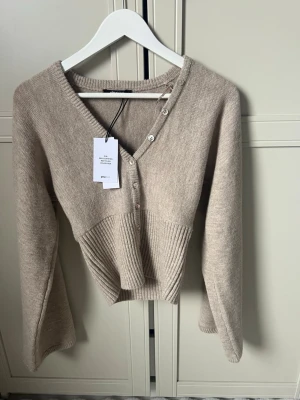 Beige stickad tröja med v-ringning - Trendig beige stickad tröja från gina tricot med djup v-ringning och dekorativa knappar framtill. Tröjan har bred ribbstickad midja och långa ärmar med lätt utsvängd form. Perfekt för dig som gillar en stilren och modern look.