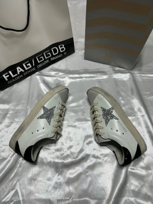 Golden Goose vita sneakers med glitterstjärna - Snygga vita sneakers från Golden Goose med silverglittrig stjärna på sidan, svarta detaljer vid häl och tå samt beige sula. Skorna har klassisk låg modell och vita skosnören. Perfekta för dig som vill sticka ut med en cool och trendig look.