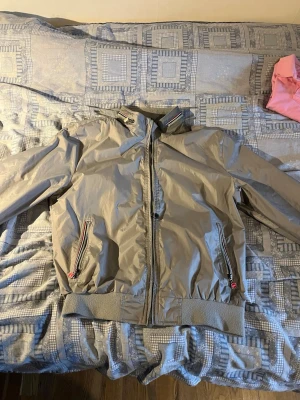 Grå vindjacka från Colmar Originals - Snygg grå vindjacka från Colmar Originals med dragkedja framtill och två fickor med dragkedja. Jackan har ribbade muddar vid ärmslut och midja samt en hög krage med diskret randig detalj. Perfekt för blåsiga dagar och enkel att matcha.