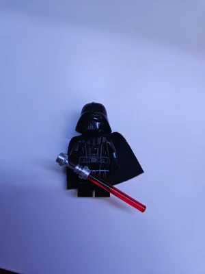 LEGO Darth Vader minifigur - LEGO minifigur föreställande Darth Vader från Star Wars. Figuren är svart med detaljerad tryck på bröstet, svart cape i tyg och en röd ljussabel i plast. Perfekt till samlingen eller för att bygga egna Star Wars-scenarion.