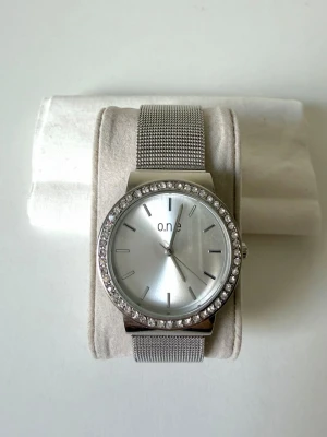 Silverfärgad klocka från o.n.e med meshband - Stilren klocka från o.n.e i silver med glittrande stenar runt urtavlan och ett snyggt mesharmband i metall. Urtavlan är minimalistisk med tydliga markeringar och en elegant känsla. Perfekt accessoar för att lyfta din outfit 🤍