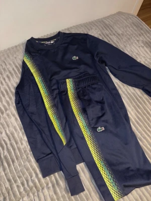 Mörkblå Lacoste sweatshirt med mönster - Mörkblå sweatshirt från Lacoste med grönt krokodilmärke på bröstet och ärmen. Snygga gula och gröna mönstrade ränder längs ena sidan och ärmen. Tröjan har rund hals och ribbade muddar. Perfekt för en sportig och trendig look.