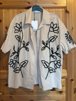 Bershka Skjorta - Stilren beige skjorta från Bershka med korta ärmar och svart broderat blommönster framtill och solmönster på ärmarna. Skjortan har klassisk krage och knäppning framtill. Perfekt för sommaren och coola detaljer. Helt ny och oanvänd. Pris kan alltid diskuteras💫💫