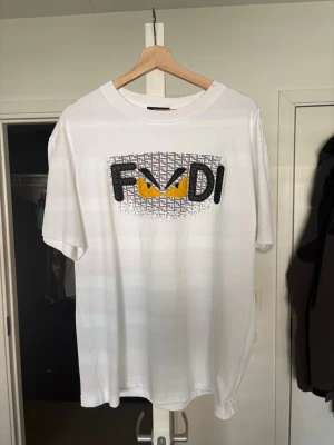 Vit Fendi t-shirt med tryck - Vit t-shirt från Fendi med stort grafiskt tryck framtill där Fendi-loggan syns tillsammans med gula ögon. Klassisk rund hals och korta ärmar. Perfekt för dig som gillar statement-plagg och exklusiva märken.