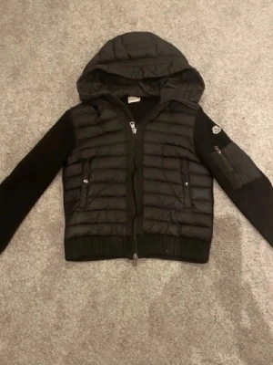 Svart pufferjacka med huva från Moncler - Snygg svart pufferjacka från Moncler med quiltad front och huva. Ärmarna är i stickat material och jackan har dragkedja framtill samt fickor med knappar. Moncler-logga på ena ärmen ger en exklusiv touch. Scam på arm och Scan på qr kod funkar 