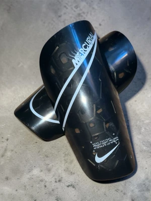 Nike Mercurial benskydd svart - Säljer ett par svarta Nike Mercurial benskydd med vit logga och mönstrad insida. Lätta och smidiga, perfekta för fotbollsträning eller match. Snygg design med hexagonmönster på insidan för extra komfort. 17cm lång och 7,5cm bred. OBS!! Små repor på dem, men funkar lika bra och luktar inget speciellt, senast använda var 4 år sedan. Skriv privat för mer bilder