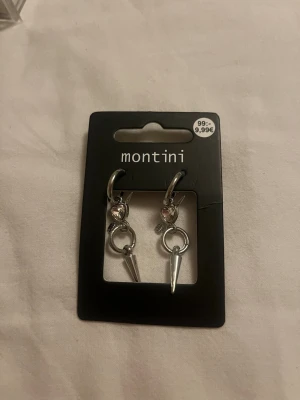 Silvriga örhängen med hjärta - Helt nya och oanvända! Snygga silvriga örhängen från Montini med hjärtformad detalj och en liten glittrig sten. Hängande ring och spetsig konformad berlock ger en cool och trendig look. Perfekta för att lyfta din stil med en edgy touch.