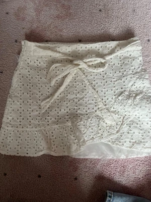 Vit omlottkjol med spets och volang - Söt vit kort omlottkjol med spetsdetaljer och volangkant. Kjolen har knytband i midjan och dragkedja i sidan. Perfekt för en somrig och fräsch look. Har sytt ihop dragkedjan så man kan inte dra i den, men det funkar om man inte har så stora höfter. Har också ett litet hål i underkjolen, men det syns inte och man stör sig inte på det! 💋