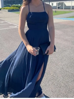 Marinblå långklänning med slits - Elegant marinblå långklänning med tunna axelband och öppen rygg. Klänningen har en hög slits på sidan och är tillverkad i ett lätt, flödande material som ger en snygg siluett. Perfekt för speciella tillfällen. Den är i storlek xs/34 och är uppsydd att passa längderna mellan 162-164 jag är 162 cm ungefär och hade klackar på bilderna. Endast använd 1 gång 