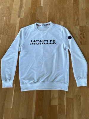 Vit Moncler sweatshirt med logga - Vit sweatshirt från Moncler med svart logotyp på bröstet och svart patch på ärmen. Tröjan har rund halsringning, långa ärmar och ribbade muddar. Perfekt för en stilren och trendig look.