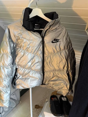 Silvrig pufferjacka från Nike - Snygg silvrig pufferjacka från Nike med svart logga på bröstet och svarta detaljer. Jackan har hög krage, dragkedja framtill och två sidofickor med dragkedja. Perfekt för kalla dagar när du vill sticka ut med en cool look.