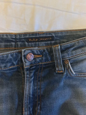 Blå jeans från Nudie Jeans Co - Säljer ett par klassiska blå jeans från Nudie Jeans Co med orangea sömmar och femficksmodell. Jeansen har en knappgylf och är tillverkade i denim av bomull. Perfekta för en avslappnad och stilren look.