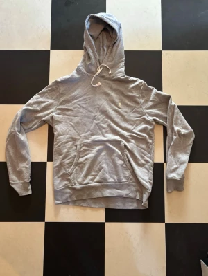 Grå hoodie från Polo Ralph Lauren - Snygg grå hoodie från Polo Ralph Lauren med klassisk broderad logga på bröstet. Tröjan har huva med dragsko och en stor magficka framtill. Tillverkad i mjuk bomullsblandning och har ribbade muddar vid ärmslut och nederkant.