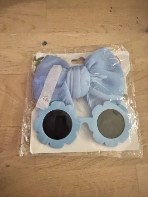 Blå blommiga solglasögon med rosett - Säljer ett par ljusblå solglasögon med blommig form på bågarna och en stor matchande rosett. Glasögonen har runda linser och är perfekta för dig som vill sticka ut med en lekfull accessoar.