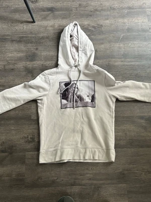 Vit hoodie med tryck framtill - Snygg vit hoodie med huva och dragsko. På bröstet finns ett svartvitt tryck med motiv. Hoodien har en avslappnad passform och är tillverkad i mjukt bomullstyg. Perfekt för en chill och stilren look.