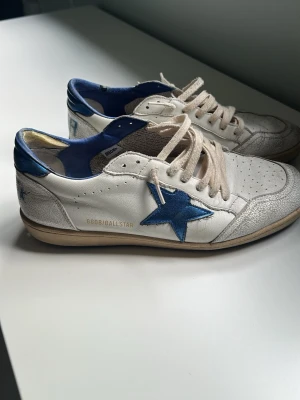 Golden Goose Ball Star vita sneakers - Golden Goose Ball Star sneakers i vitt läder med blå detaljer och stjärna på sidan. Skorna har perforerad tå, beige snörning och blå hälkappa. Snygg retrostil med slitna detaljer och gummisula. Perfekta för dig som gillar streetwear och unik design. Pris går att förhandlas. Passar 45