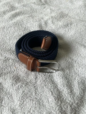 Mörkblått flätat bälte med skinn - Snyggt mörkblått flätat bälte med bruna skinn-detaljer och silverfärgat spänne. Perfekt accessoar för att lyfta din outfit med en klassisk touch. Bältet har en stilren design och passar till många olika looks.