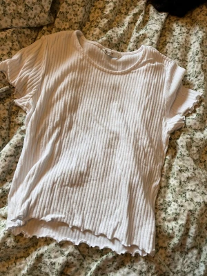Vit ribbad topp från ONLY - Vit ribbad topp från ONLY med korta ärmar och rund halsringning. Toppen har en avslappnad passform och vågig kant nertill för en trendig look. Perfekt att styla med jeans eller kjol för en chill vibe.
